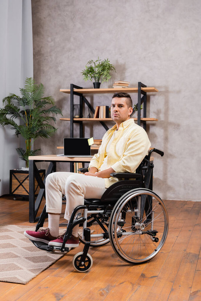 jeune homme handicapé en fauteuil roulant regardant la caméra près de l'ordinateur portable avec écran blanc sur le bureau - Photo, image