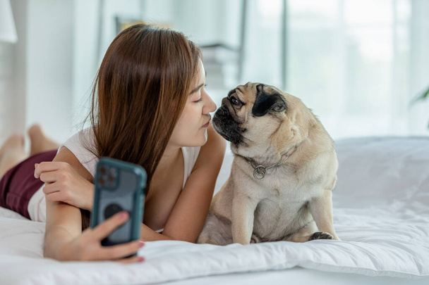 Beautiful Asian young woman take a photo selfie bu mobile phone with her cute dog pug breed in cozy bedroom,Happiness girl playing with dog to take a rest at home in holiday - Fotografie, Obrázek