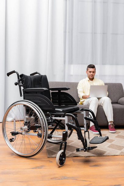 handicapped man working on sofa with laptop near wheelchair on foreground - Foto, Imagem