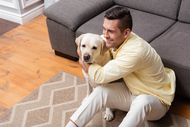 Blick aus der Vogelperspektive auf einen fröhlichen Mann, der Labrador-Hund kuschelt, während er auf der Couch neben dem Sofa sitzt - Foto, Bild