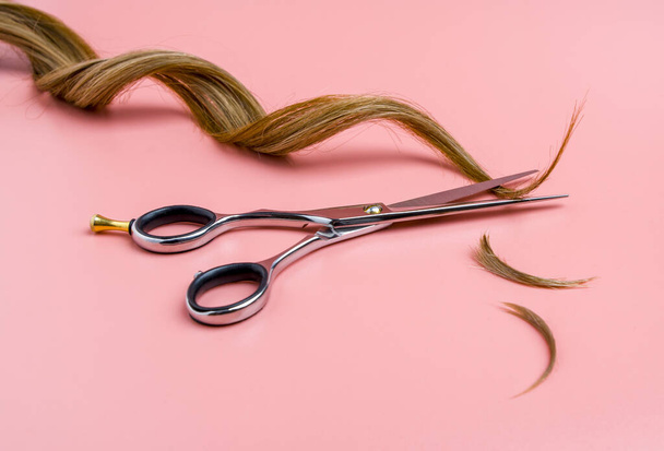 Open kappers schaar met een streng van golvend haar op roze achtergrond, professionele schaar en geslepen haarranden - Foto, afbeelding