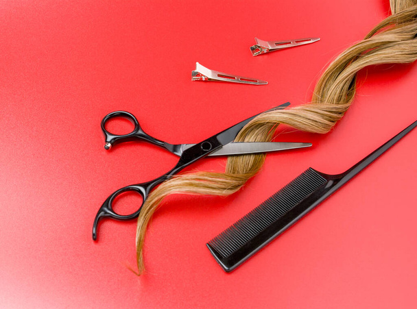 Otevřené kadeřnické nůžky s pramenem kudrnatých umělých vlasů na červeném pozadí, profesionální nůžky, černý hřeben, kovové klipy a ostříhané okraje vlasů - Fotografie, Obrázek