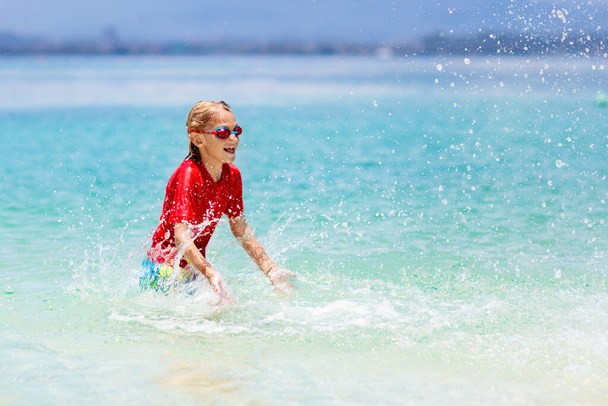 Niños jugando en la playa tropical. Agua salpicada de diversión. Un niño salpicando olas marinas. Los niños nadan y juegan en vacaciones familiares de verano. Juguete de arena y agua, protección solar para niños pequeños.  - Foto, imagen
