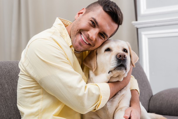 zufriedener Mann lächelt in die Kamera, während er zu Hause Labrador knuddelt - Foto, Bild