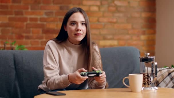 Ocio doméstico. Mujer joven jugando videojuegos en casa, sentada en un sofá con joystick y sintiéndose emocionada en casa - Imágenes, Vídeo