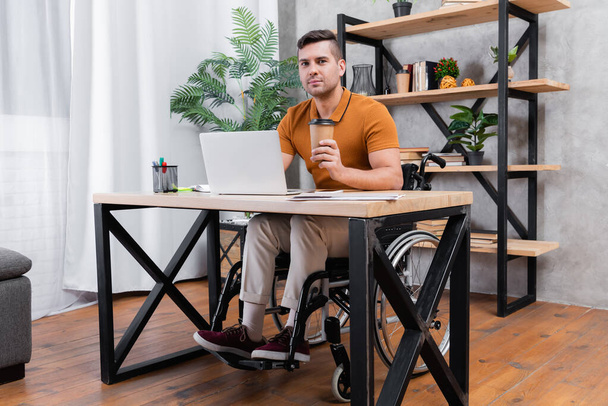 niepełnosprawny mężczyzna trzyma kawę, aby przejść podczas pracy w pobliżu laptopa w domu - Zdjęcie, obraz