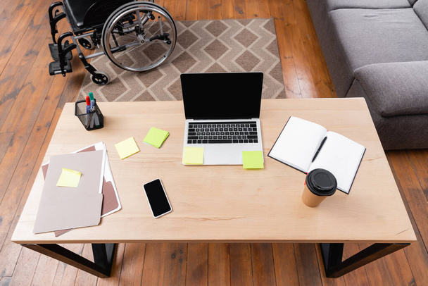 high angle view of desk with gadgets, documents and takeaway drink near wheelchair - Photo, Image