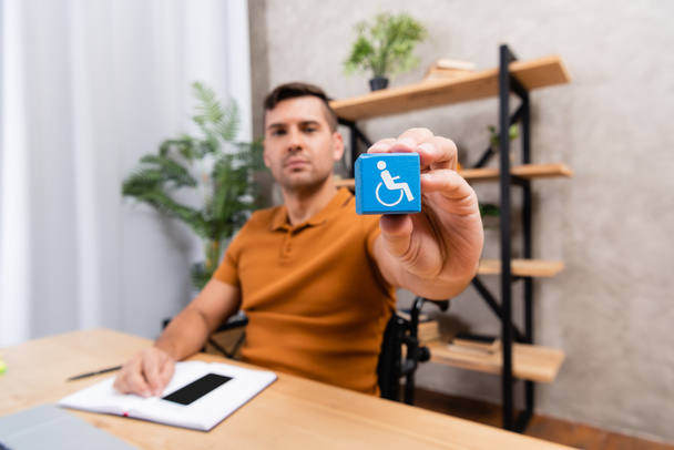 jeune homme montrant un cube avec signe de handicap alors qu'il était assis au bureau à la maison, fond flou - Photo, image