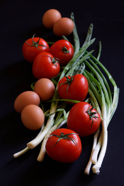 Φρέσκες κόκκινες ντομάτες, πράσινα κρεμμύδια και αυγά κοτόπουλου βρίσκονται σε μια σκοτεινή επιφάνεια. Ρουστίκ έννοια πρωινό, συστατικά σαλάτας και υγιεινά προϊόντα. Κοντινό πλάνο - Φωτογραφία, εικόνα