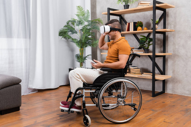 vammainen mies hymyilee pelatessaan vr kuulokkeissa pyörätuolissa kotona - Valokuva, kuva