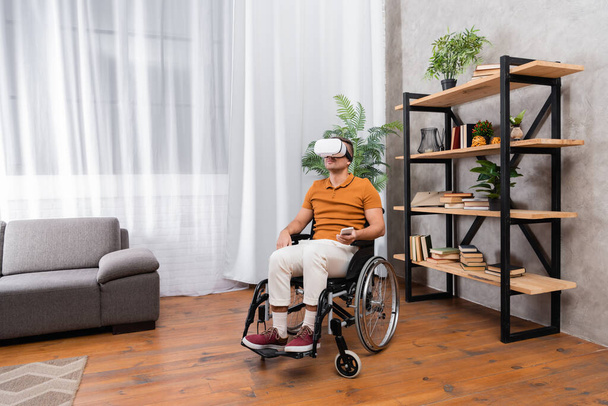 Evde tekerlekli sandalyede oturan engelli adam kulaklıkla oyun oynuyor. - Fotoğraf, Görsel