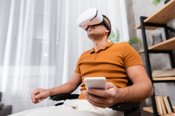 low angle view of handicapped man holding smartphone while gaming in vr headset - Photo, image