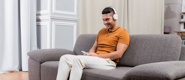 uomo sorridente seduto sul divano, chatta su smartphone e ascolta musica in cuffia, banner - Foto, immagini