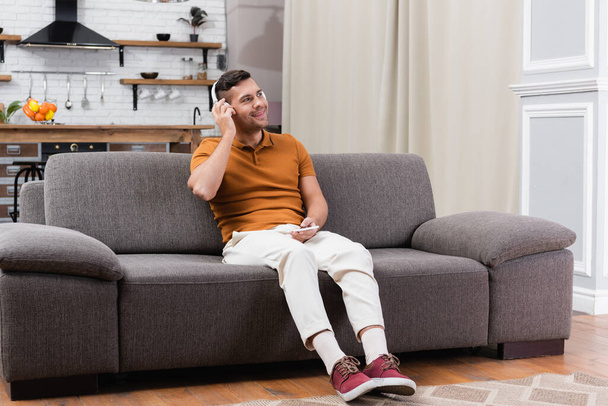 joyful man holding smartphone while listening music in headphones on sofa - Foto, Imagem