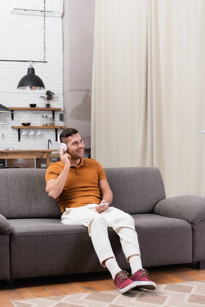 smiling man listening music in headphones while sitting on couch with smartphone - Photo, Image