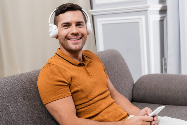 cheerful man smiling at camera while listening music in wireless headphones - Фото, изображение