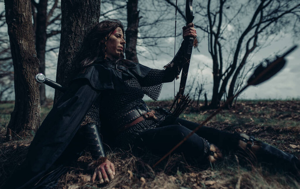 Woman in chain mail in image of medieval warrior sits and rests near tree among forest. - Photo, Image