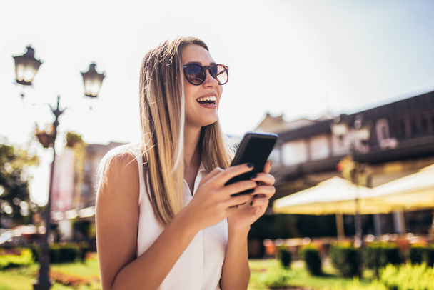 Prachtige mooie jonge vrouw met blond haar messaging op de smartphone op de stad straat achtergrond - Foto, afbeelding