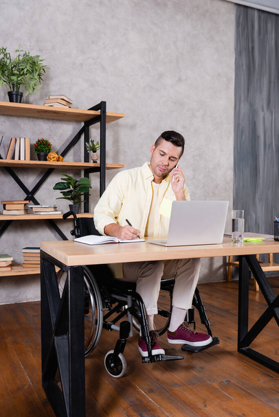 disabled man writing in notebook and talking on mobile phone while working at home - Foto, Imagem