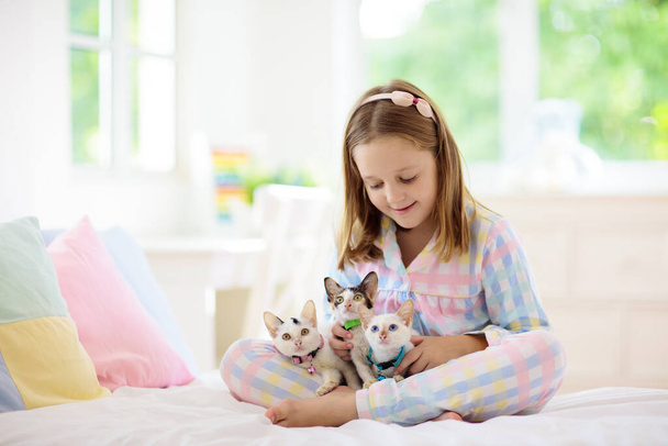 Kind spelen met baby kat op bed in witte slaapkamer. Een kind met een wit poesje. Klein meisje in pyjama met schattig huisdier thuis. Kinderen spelen met katten. Kinderen en huisdieren huisdieren. - Foto, afbeelding