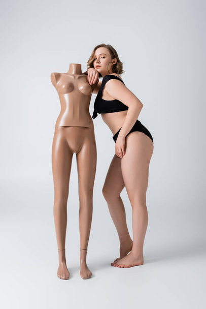 plná délka nadváhy a bosá žena v plavkách opírající se o plastovou figurínu na bílém  - Fotografie, Obrázek