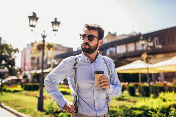 Красивий молодий чоловік у розумному повсякденному одязі стоїть на вулиці, тримаючи чашку кави
 - Фото, зображення