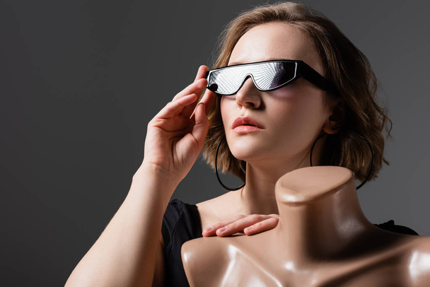 overweight young woman adjusting sunglasses and posing with plastic mannequin isolated on grey - Фото, зображення