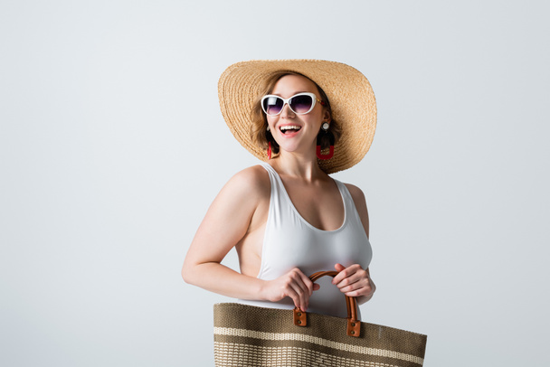 femme en surpoids et joyeuse en chapeau de paille, lunettes de soleil et maillot de bain debout avec sac isolé sur blanc - Photo, image