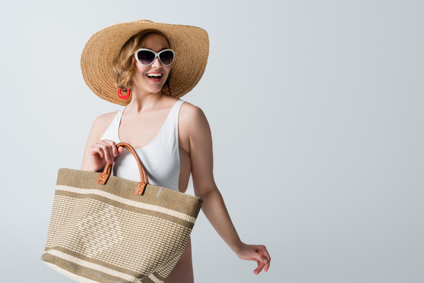 overweight and joyful woman in straw hat, sunglasses and swimsuit holding bag isolated on white - 写真・画像