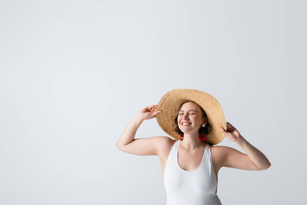 overweight and pleased woman in earrings and swimsuit adjusting straw hat isolated on white - 写真・画像