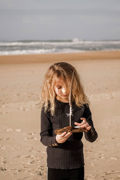 Kız kumlu sahilde elinde odun ve martı tüyüyle elinde oyuncaklarla bekliyor. Arka planda plaj manzarası var. Yüksek kalite fotoğraf - Fotoğraf, Görsel