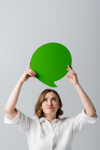 displeased overweight woman in white shirt holding green speech bubble above head isolated on grey  - Foto, afbeelding