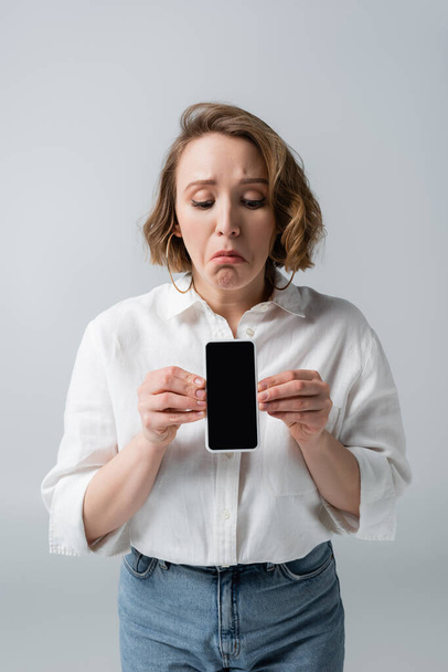 triste joven mujer con sobrepeso sosteniendo teléfono inteligente con pantalla en blanco aislado en gris - Foto, imagen