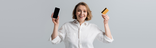 happy plus size woman holding credit card and smartphone isolated on grey, banner - Photo, Image