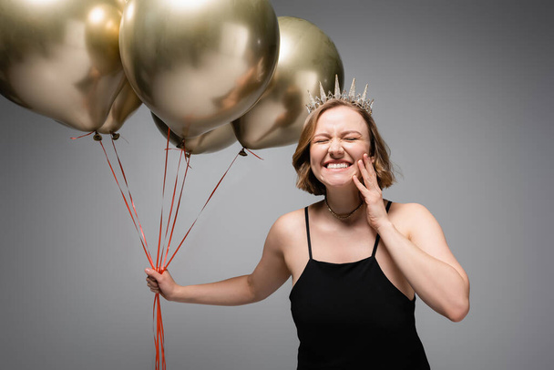 happy plus size woman in black slip dress and crown holding golden balloons on grey  - Photo, Image