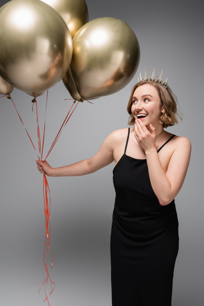 excited plus size woman in black slip dress and crown holding golden balloons on grey  - Foto, Imagem