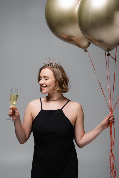 happy plus size woman in black slip dress and crown holding golden balloons and champagne isolated on grey  - Foto, Imagem
