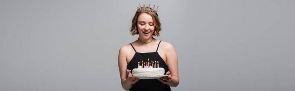 feliz mulher plus size em vestido de deslizamento e coroa segurando bolo de aniversário isolado em cinza, banner - Foto, Imagem