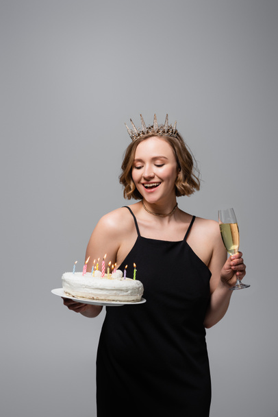 happy plus size woman in slip dress and crown holding birthday cake and glass of champagne isolated on grey  - Fotó, kép