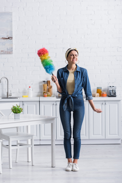 Enojado ama de casa sosteniendo polvo cepillo en cocina  - Foto, Imagen