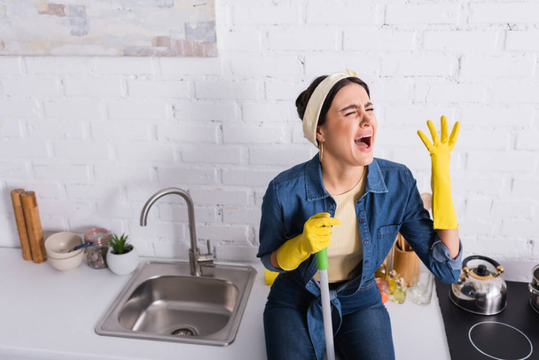 Displeased housewife with mop sitting on kitchen worktop  - 写真・画像