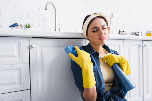 Displeased housewife in rubber gloves holding sponge and detergent in kitchen  - Photo, Image