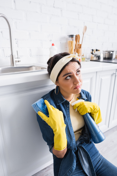 Pensive housewife with sponge and detergent near kitchen worktop  - Фото, изображение