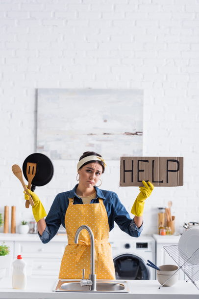 Θλιβερή νοικοκυρά κρατώντας πλακάτ με βοήθεια γράμματα και σκεύος στην κουζίνα  - Φωτογραφία, εικόνα