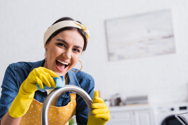 Χαρούμενη γυναίκα που καθαρίζει βρύση κουζίνας με σφουγγάρι  - Φωτογραφία, εικόνα