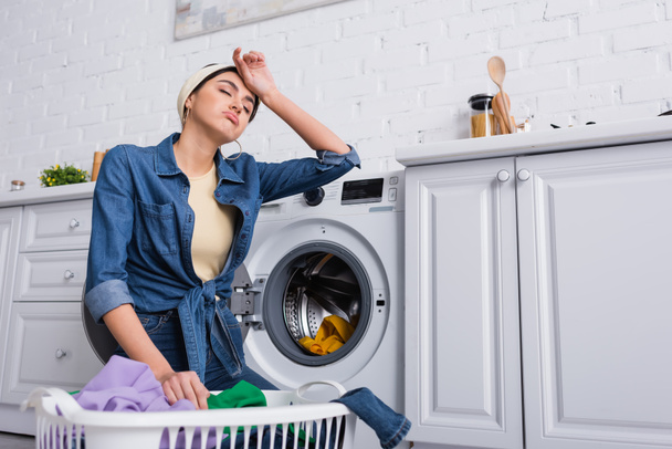 Femme au foyer fatiguée près du panier avec des vêtements et lave-linge dans la cuisine  - Photo, image