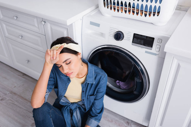 Overhead view of exhausted housewife sitting near washing machine in kitchen  - Foto, Imagen
