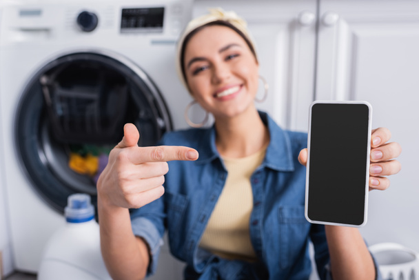 Blurred housewife pointing at smartphone near washing machine  - 写真・画像