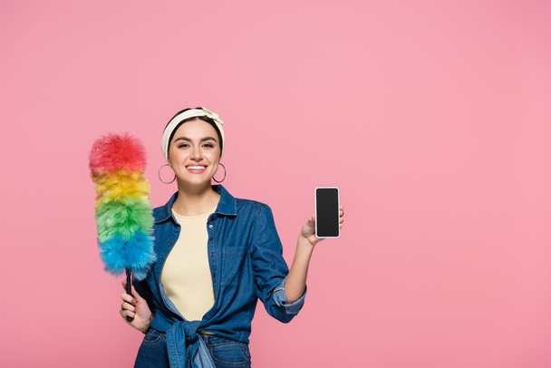 Femme au foyer souriante avec brosse à poussière tenant téléphone portable avec écran blanc isolé sur rose - Photo, image