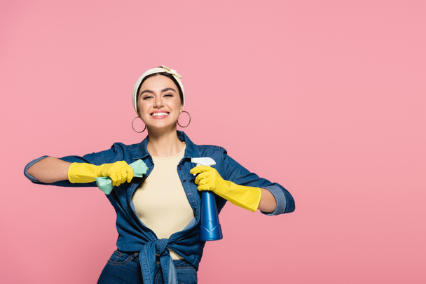 Позитивна жінка в гумових рукавичках тримає пральний порошок і ганчірку ізольовано на рожевому
 - Фото, зображення